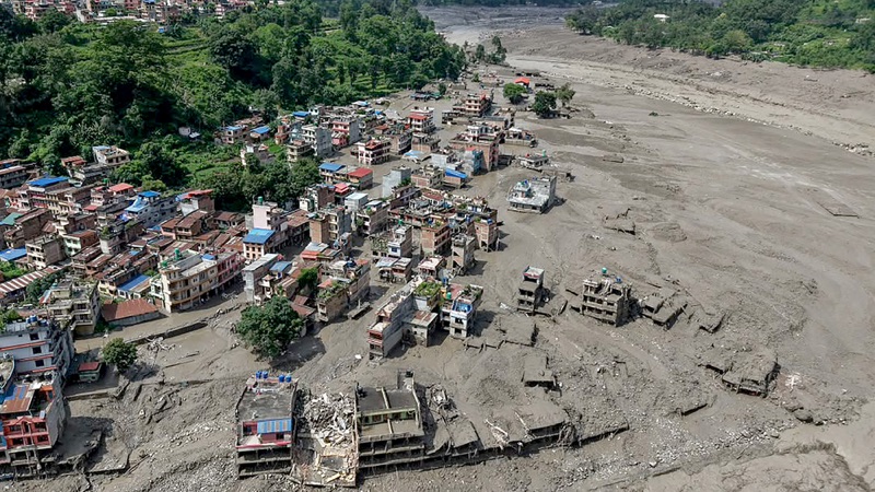 Aerial view of a riverside town where a wide expanse of grey mud and debris has cut through the settlement. Several multi-storey concrete buildings stand gutted and partly collapsed in the mud, while denser rows of houses and green hillside remain to one side.